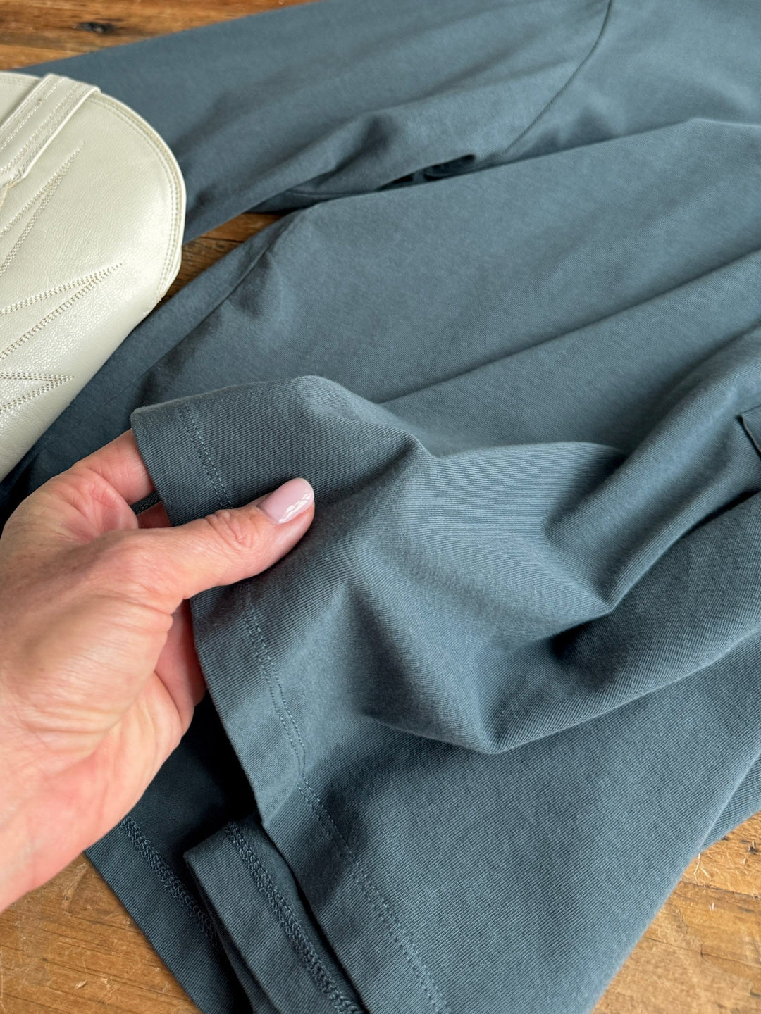 A hand with light pink nail polish holds the hem of the Colville Slate Basic Top by Savannah Sevens western life{&}style, a dark gray long sleeve piece, on a wooden surface. A cream-colored item with stitched details is partially visible on the left.