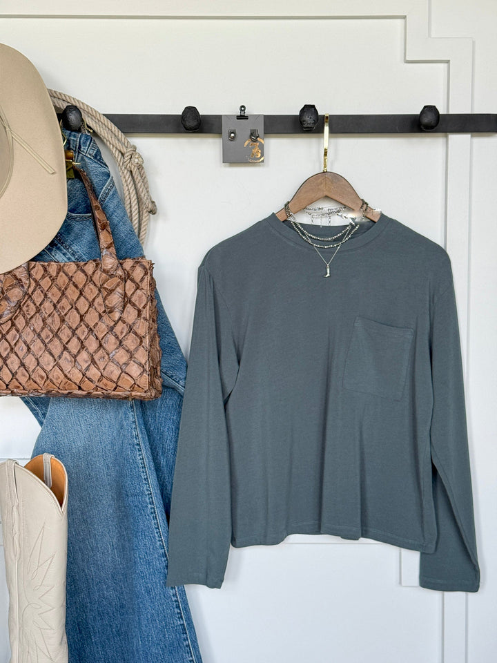 A Colville Slate Basic Top by Savannah Sevens western life{&}style on a hanger, paired with layered necklaces, blue jeans, a woven brown handbag, a tan hat, and cream cowboy boots displayed on black wall hooks.
