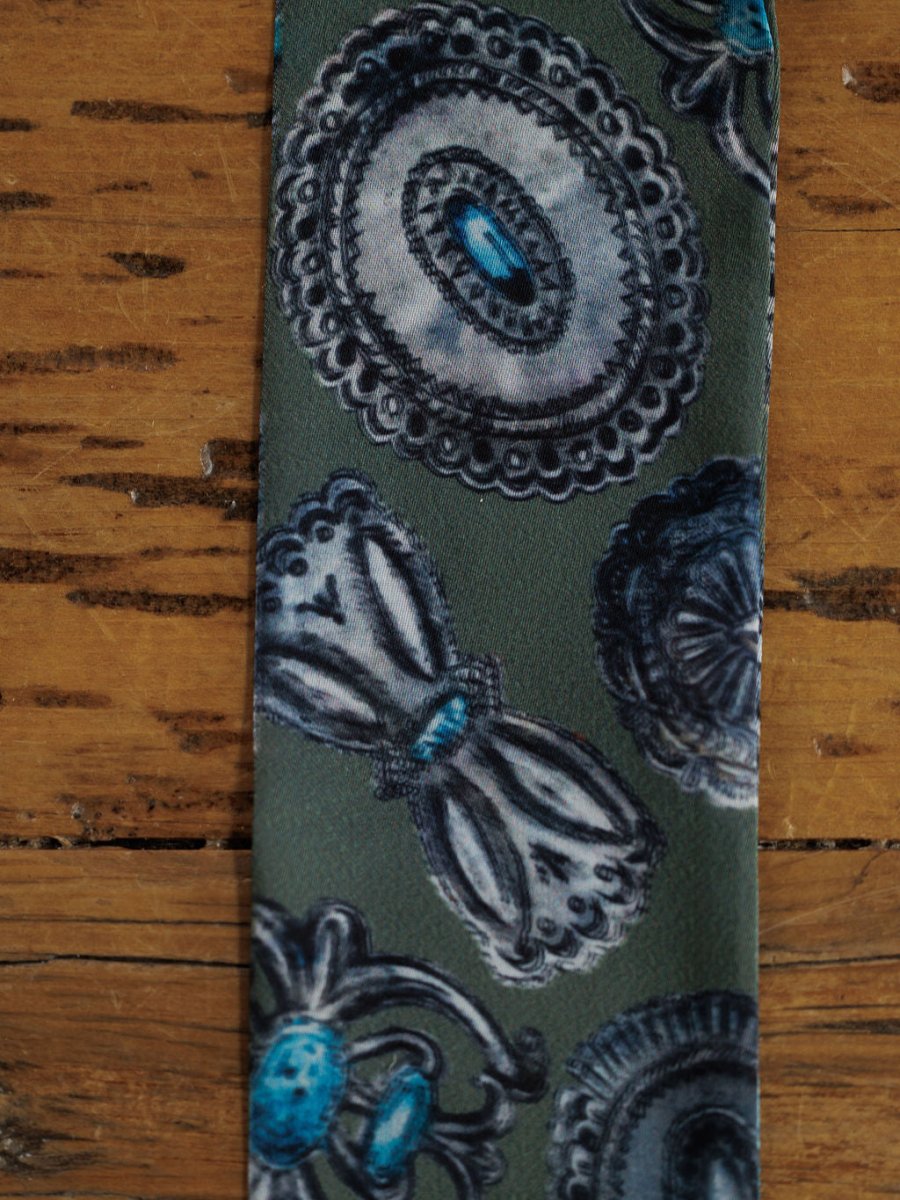 A close-up of the Concho Twilly {Sage} satin tie scarf by Savannah Sevens western life{&}style, featuring blue and gray circular and bow-like designs on a green background, displayed on a rustic wooden surface.