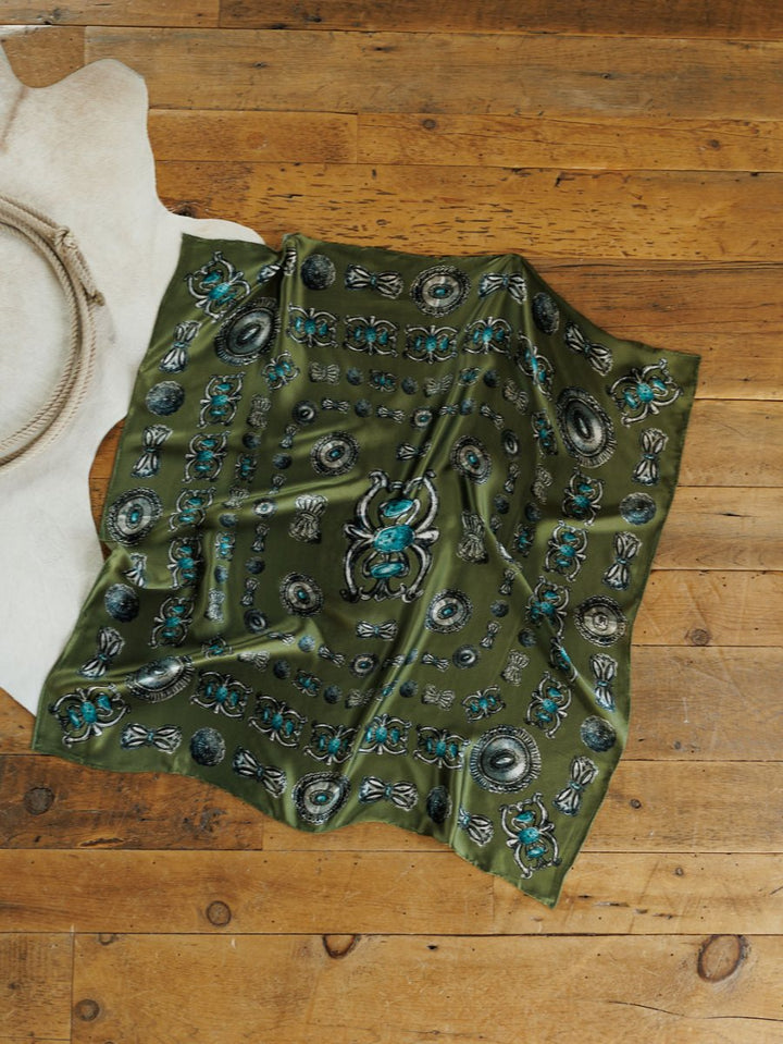 The Concho Wild Rag by Savannah Sevens western life{&}style, a green silk scarf with blue, black, and white patterns plus hand-sewn trim, is displayed on a wooden floor beside white animal hide and a coiled rope.