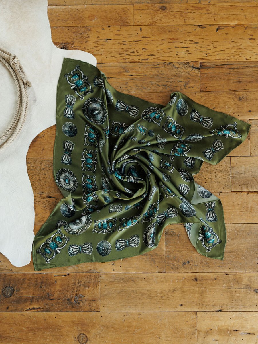 The Concho Wild Rag from Savannah Sevens western life{&}style, featuring green silk with blue and black patterns and a hand-sewn trim, is displayed on a wooden floor beside a coiled rope and white cowhide rug.