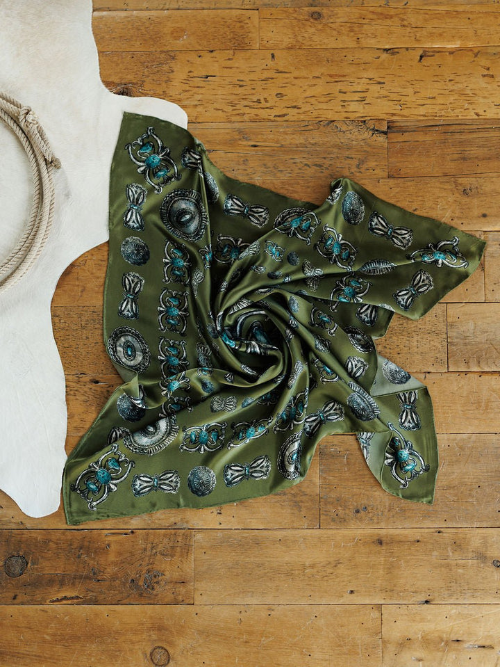 The Concho Wild Rag from Savannah Sevens western life{&}style, featuring green silk with blue and black patterns and a hand-sewn trim, is displayed on a wooden floor beside a coiled rope and white cowhide rug.