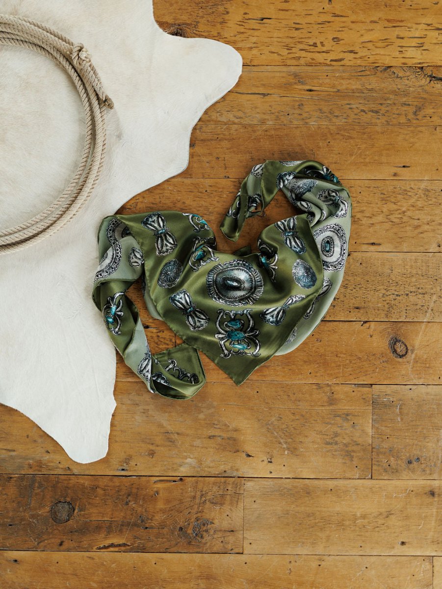 The Concho Wild Rag by Savannah Sevens western life{&}style, a green patterned silk scarf with hand-sewn trim, is styled in a heart shape on wood near a white fur rug with coiled rope for distinctive western flair.