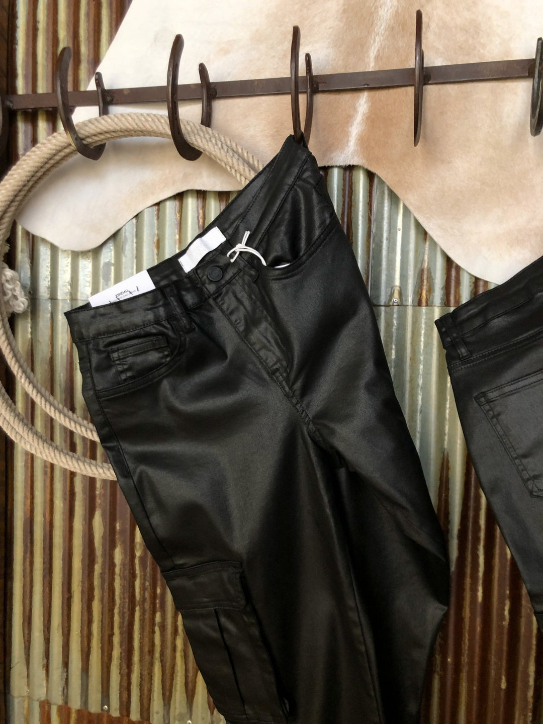 The Conrad High Rise 90's Straight Leg Jean by Savannah Sevens western life{&}style hangs on a rustic metal hook rack, with animal hide in the background against corrugated metal.