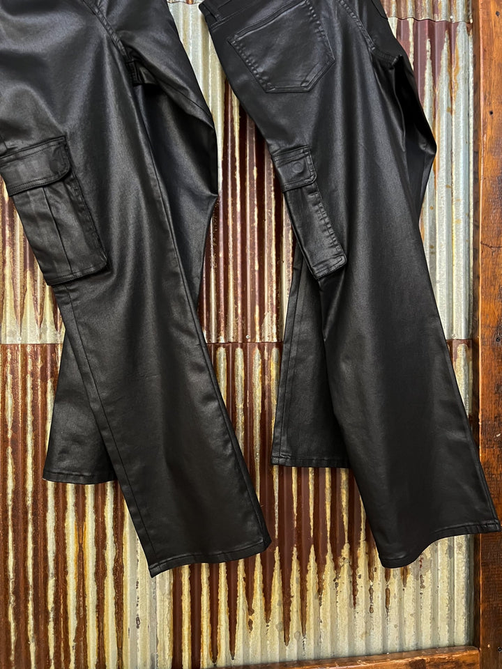 Two pairs of The Conrad High Rise 90's Straight Leg Jean by Savannah Sevens western life{&}style, featuring black faux leather and cargo pockets, hang side by side against a rusty corrugated metal wall, with a wooden frame on the right.