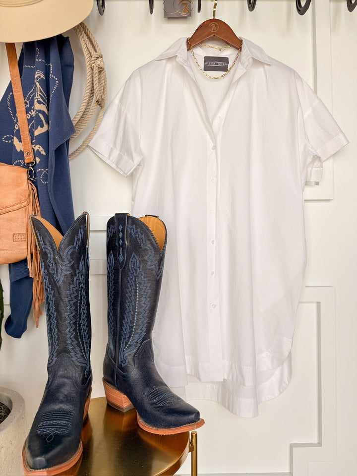 The Conrad White Cotton Button Up Dress by Savannah Sevens western life{&}style hangs next to black cowboy boots with blue stitching, a tan hat, a rope, a blue cowboy print scarf, and a small tan purse.