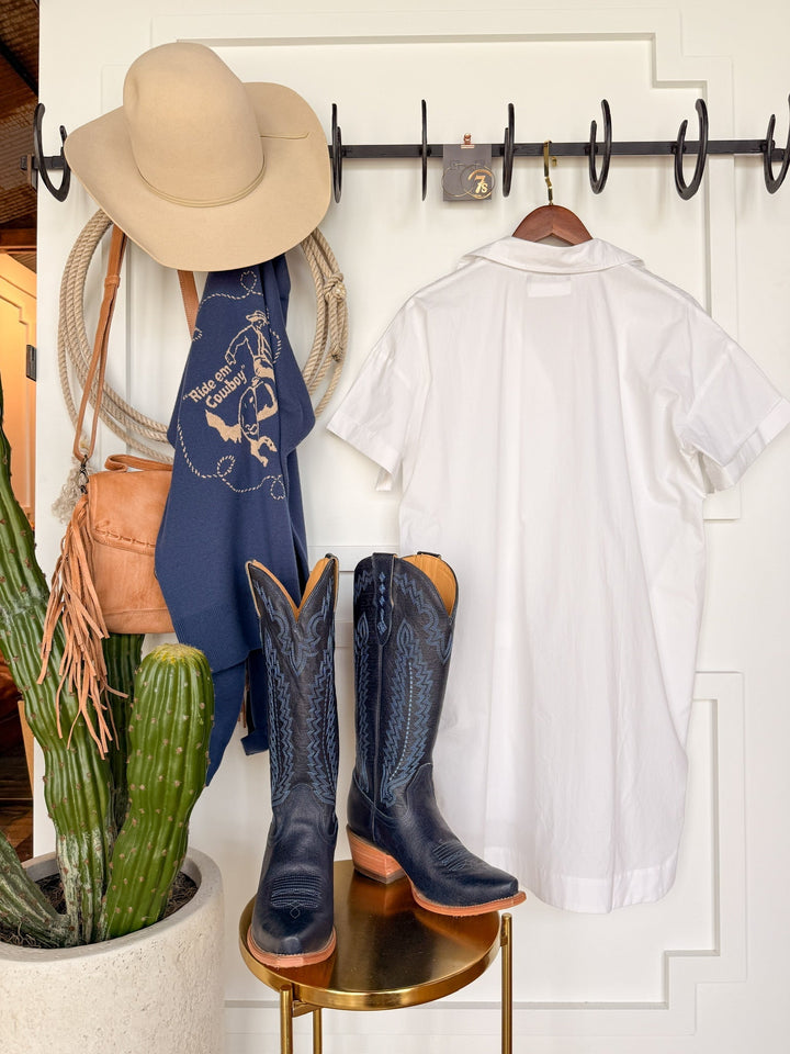 The Conrad White Cotton Button Up Dress by Savannah Sevens western life{&}style hangs above black cowboy boots, alongside a beige wide-brim hat, blue bandana, tan fringe bag, and a green potted cactus for a Western boho look.