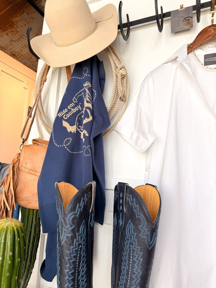 A beige cowboy hat and blue "Ride em Cowboy!" sweater hang above the Conrad White Cotton Button Up Dress by Savannah Sevens western life{&}style. Black and blue cowboy boots stand by a green cactus, with a tan bag also hanging on the hooks.