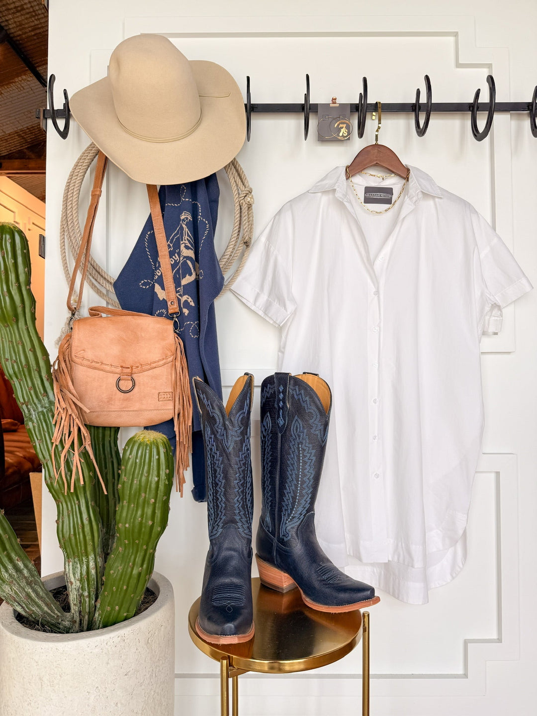 The Conrad White Cotton Button Up Dress by Savannah Sevens western life{&}style hangs with a beige wide-brim hat, tan fringe purse, blue patterned scarf, and black cowboy boots above a cactus against a white wall and gold side table.