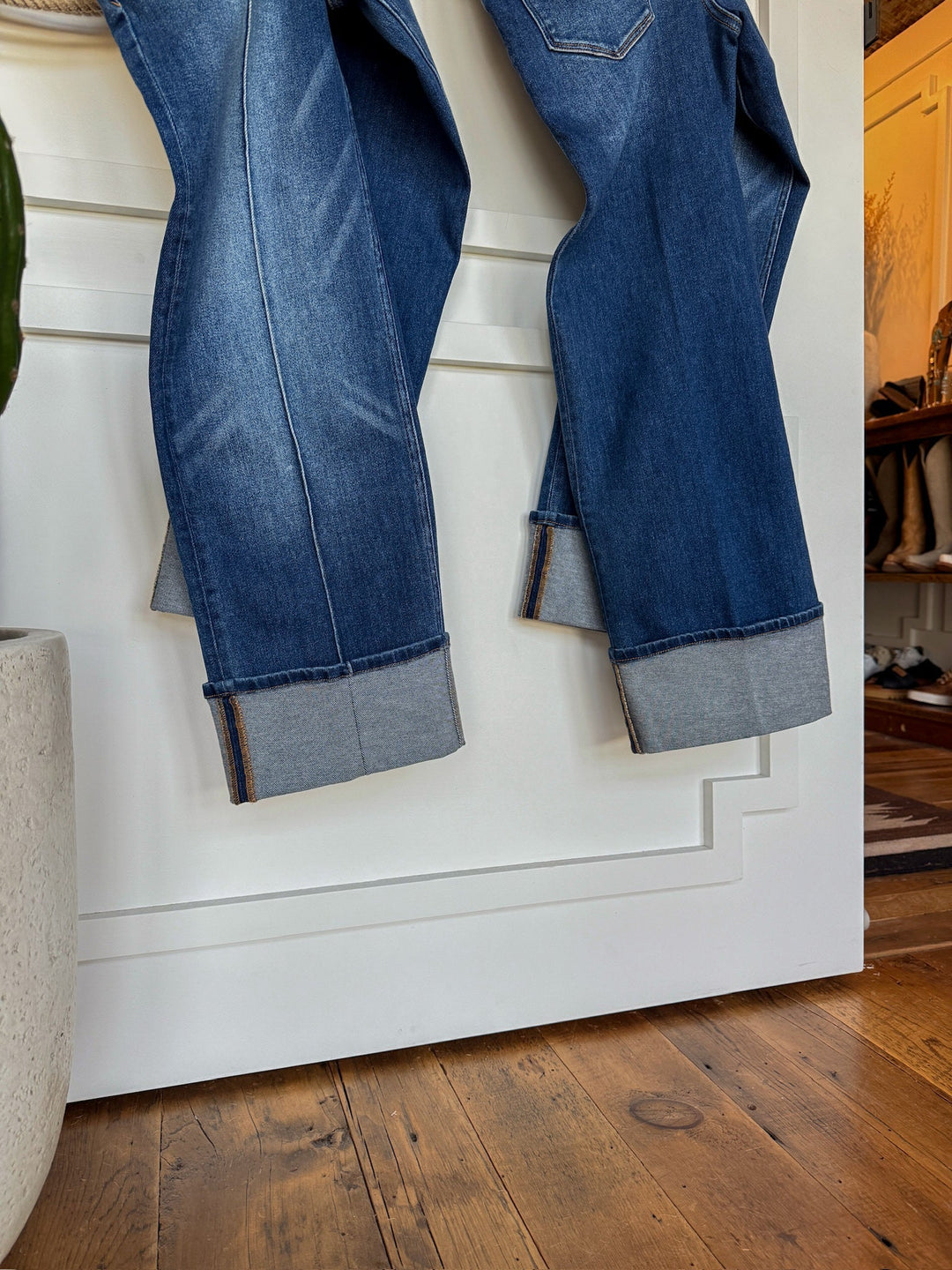Two pairs of Savannah Sevens western life{&}style The Conway High Rise Slim Wide Leg Jeans in blue, featuring cuffed hems, hang side by side on a white paneled door. A wooden floor and part of a plant pot are seen in the foreground.