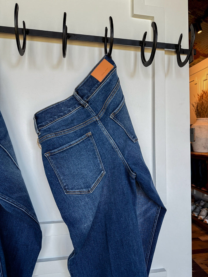 A pair of Savannah Sevens western life{&}style The Conway High Rise Slim Wide Leg Jeans hangs from a black wall-mounted hook rack against a white paneled wall, showing the back pockets and leather waistband patch.