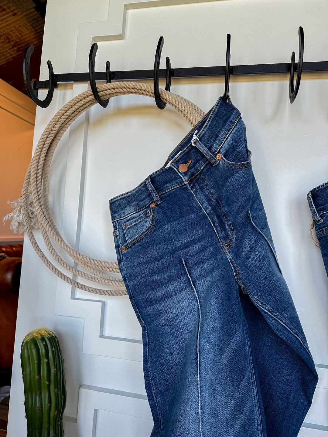 A pair of Savannah Sevens western life{&}style The Conway High Rise Slim Wide Leg Jeans hangs on a black metal hook rack mounted on a white wall, with a coiled rope and a green cactus nearby.