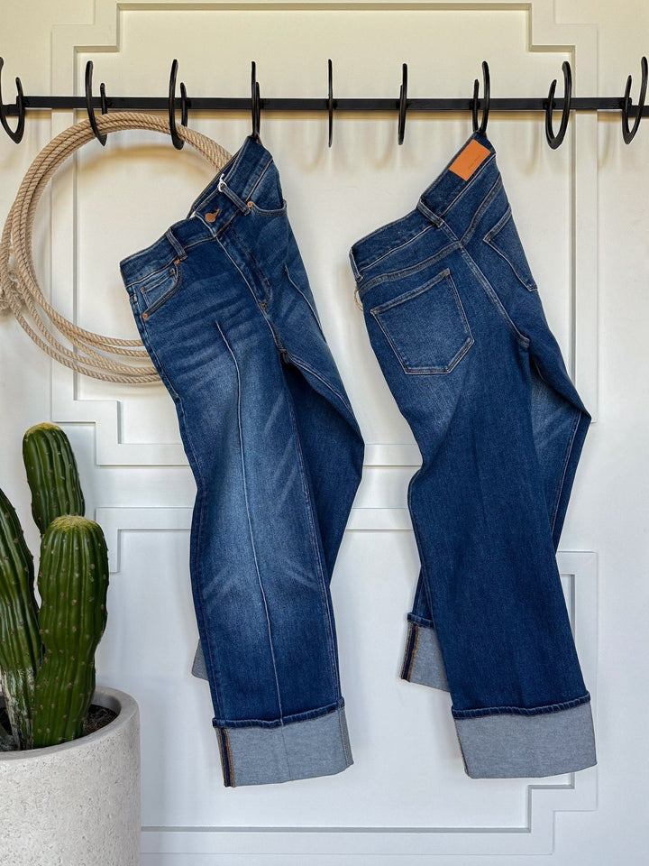 Two pairs of Savannah Sevens western life{&}style’s The Conway High Rise Slim Wide Leg Jeans with rolled cuffs hang on black hooks by a white paneled door, near a coiled rope and potted cactus, highlighting their ultra high rise style.