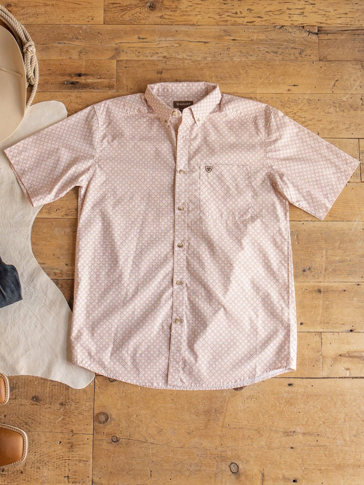 The Copeland Ariat Men's Southwest Button Up from Savannah Sevens western life{&}style, featuring a light pink hue and subtle geometric pattern, is displayed flat on a wooden floor beside a white rug and tan boots, embodying modern southwestern style.