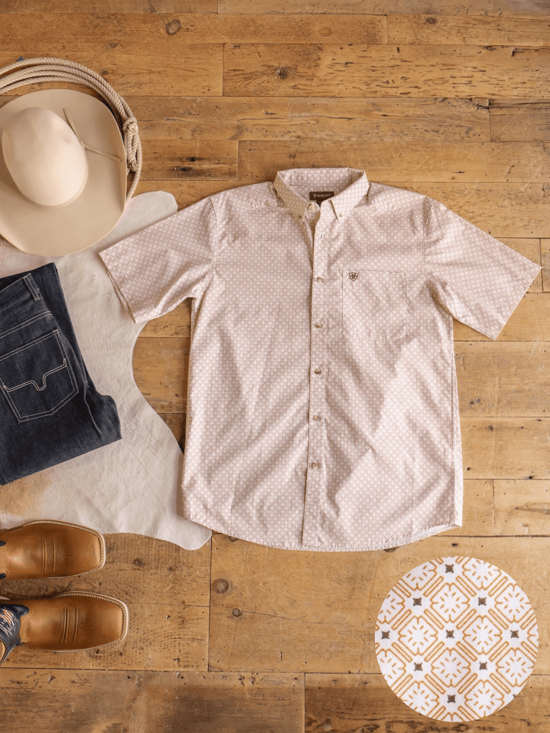 The Copeland Ariat Men's Southwest Button Up by Savannah Sevens western life{&}style, featuring short sleeves in light pink, is neatly displayed on a wooden floor alongside jeans, brown shoes, a cowboy hat, a cowhide rug, and a round decorative tile with a geometric pattern.