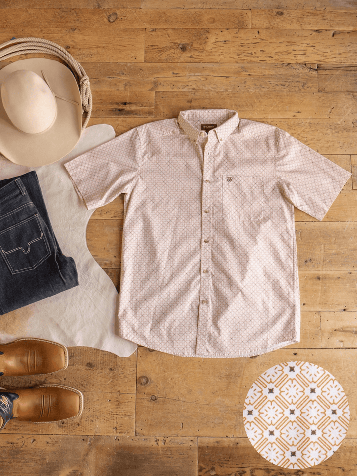The Copeland Ariat Men's Southwest Button Up by Savannah Sevens western life{&}style, featuring short sleeves in light pink, is neatly displayed on a wooden floor alongside jeans, brown shoes, a cowboy hat, a cowhide rug, and a round decorative tile with a geometric pattern.