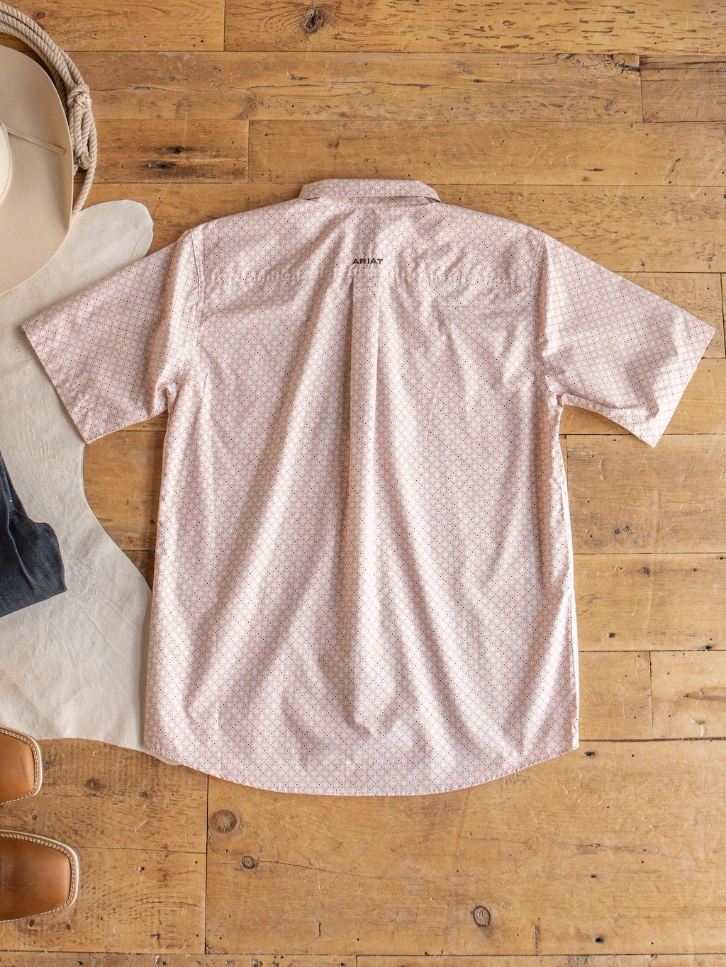 The Copeland Ariat Men's Southwest Button Up from Savannah Sevens western life{&}style is displayed laid flat on a wooden floor in a light pink pattern, shown from the back and styled next to jeans, a hat, and brown shoes.