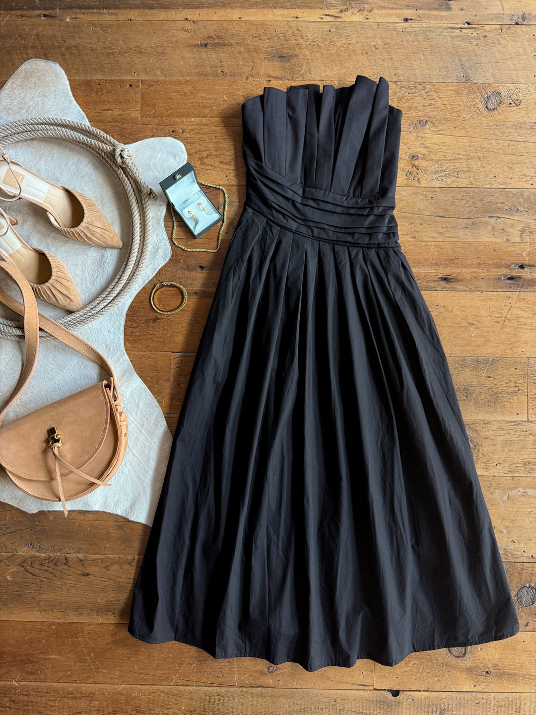 The Coppell Black Strapless Pleat Dress by Savannah Sevens western life{&}style is laid out on a wooden floor, styled with tan heels, a tan purse, a bracelet, and a small blue perfume bottle beside it.
