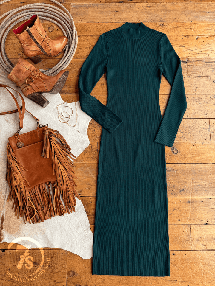 The Coppell Emerald Ribbed Long Sleeve Dress by Savannah Sevens western life{&}style is styled on a wooden floor with brown ankle boots, a fringed crossbody bag, gold hoop earrings, and coiled rope atop a white cowhide rug.
