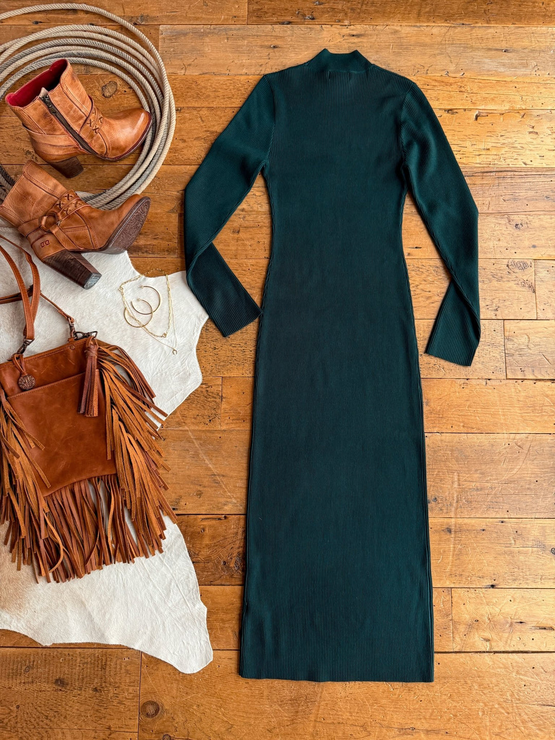 The Coppell Emerald Ribbed Long Sleeve Dress by Savannah Sevens western life{&}style is displayed on a wooden floor with tan cowboy boots, a fringed brown suede handbag, hoop earrings, and a gold bracelet.