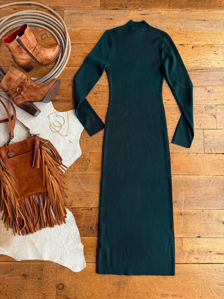The Coppell Emerald Ribbed Long Sleeve Dress by Savannah Sevens western life{&}style is displayed on a wooden floor with tan cowboy boots, a fringed brown suede handbag, hoop earrings, and a gold bracelet.
