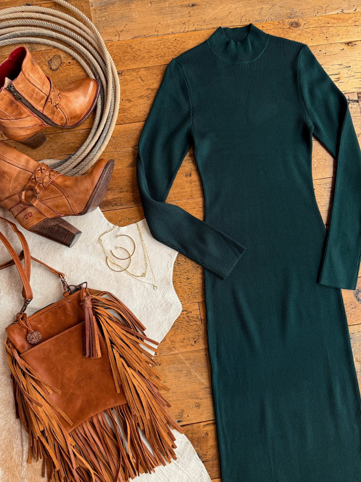 Flat lay of the Coppell Emerald Ribbed Long Sleeve Dress by Savannah Sevens western life{&}style, paired with brown heeled ankle boots, a brown fringe handbag, and gold jewelry on a wooden floor with a light cowhide rug.
