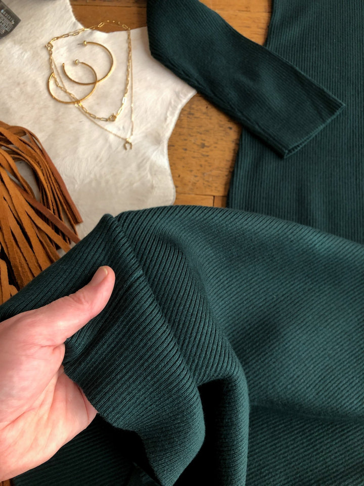 A hand holds the Coppell Emerald Ribbed Long Sleeve Dress by Savannah Sevens western life{&}style on a wooden table, shown with gold jewelry, tan fringe, and soft white fabric.