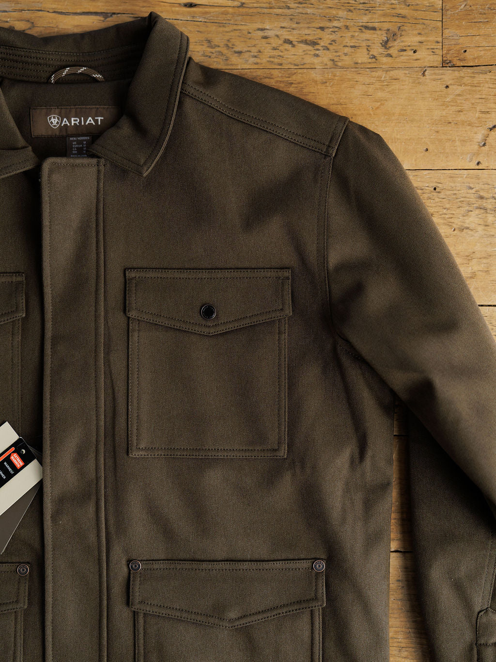 The Cornell Ariat Men's Insulated Jacket by Savannah Sevens western life{&}style is shown in olive green, laid flat with buttoned chest and lower pockets, visible collar, clothing tag on the left, and a wind and water resistant design for all-weather wear.