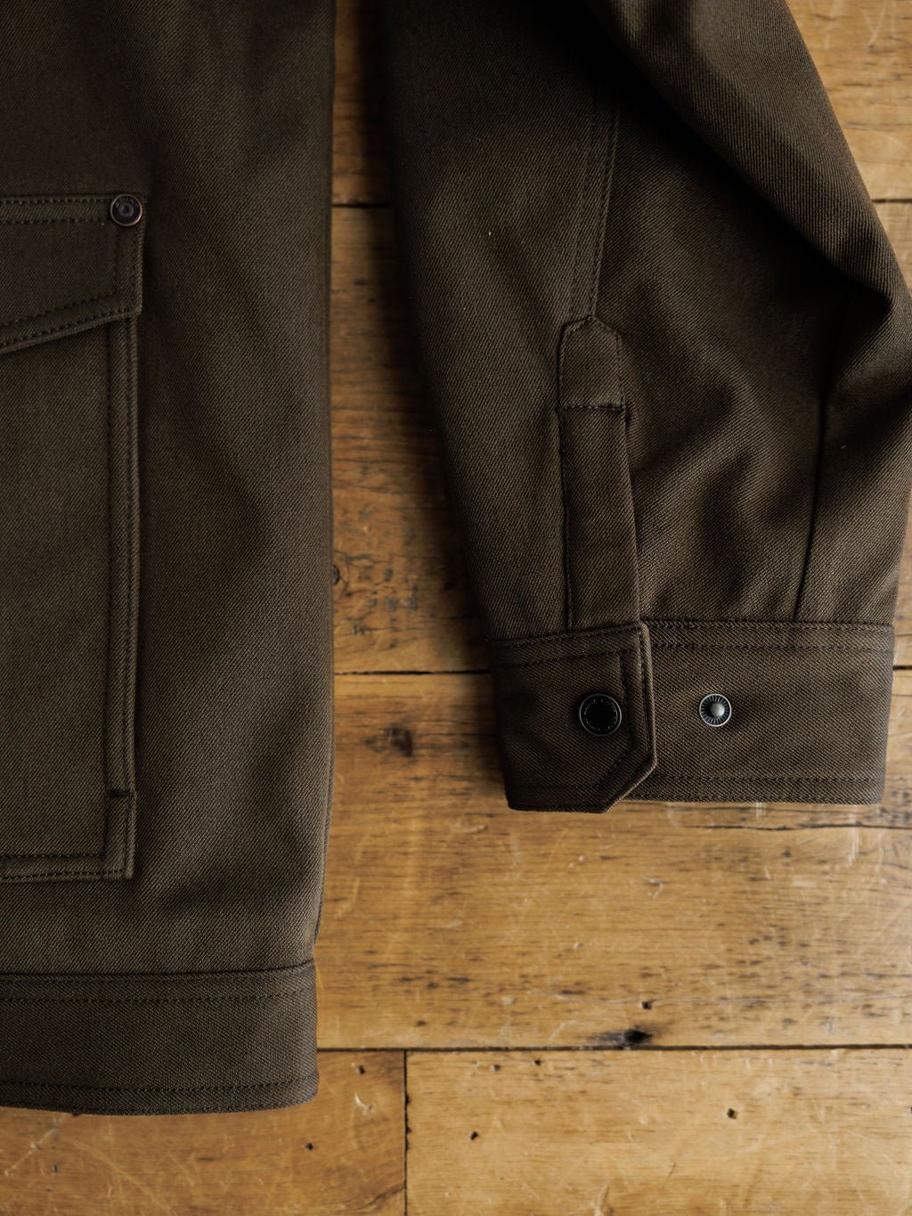 A close-up of the Cornell Ariat Men's Insulated Jacket by Savannah Sevens western life{&}style in dark olive green, highlighting the sleeve's buttoned cuff and detailed stitching on its wind and water resistant fabric, laid on rustic wood.