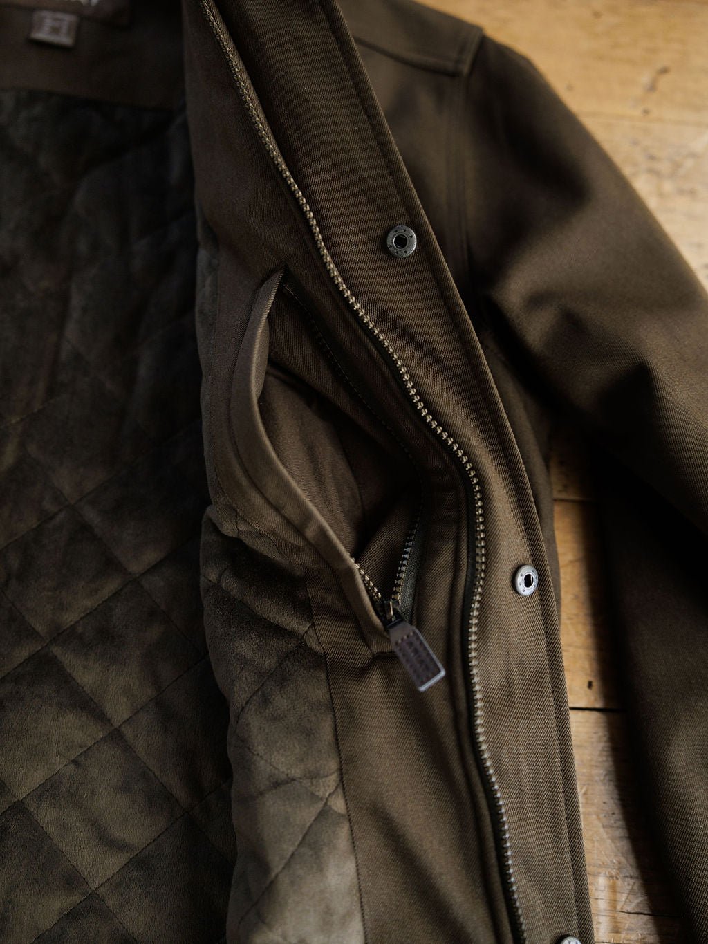 A close-up of the Cornell Ariat Men's Insulated Jacket by Savannah Sevens western life{&}style in olive green on a wooden surface, showing its quilted lining, wind and water resistant fabric, zippered inner pocket, and outer buttons.
