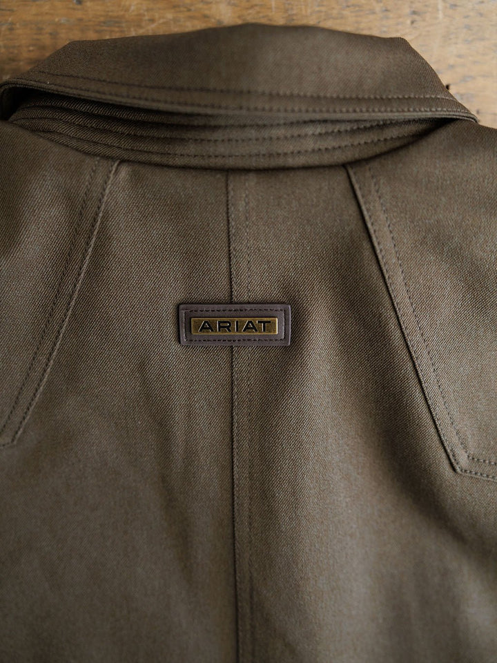 Close-up of the back of a Cornell Ariat Men's Insulated Jacket by Savannah Sevens western life{&}style, showing a stitched brand label near the collar on brown fabric. The jacket is wind and water resistant, with a wooden background.