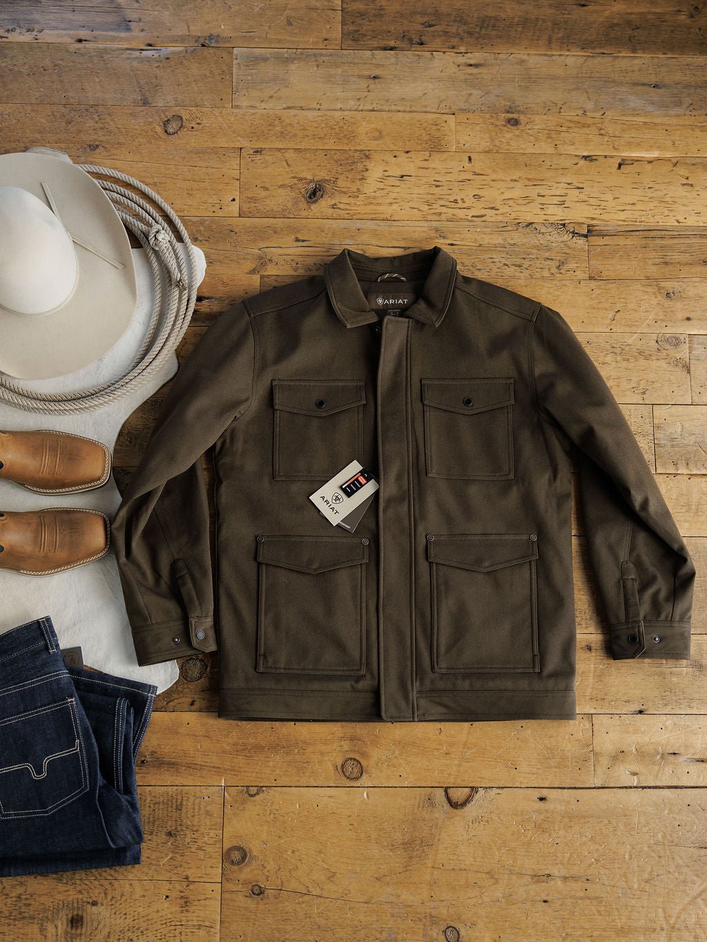 A Cornell Ariat Men’s Insulated Jacket from Savannah Sevens western life{&}style, in olive green, is laid on a wooden floor beside tan boots, folded denim jeans, a coiled rope, and a white cowboy hat. The jacket features wind and water resistance.