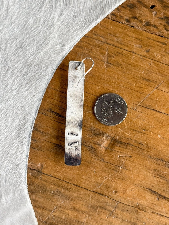 The Corrales Earrings by Savannah Sevens western life{&}style, rectangular and marked STERLING, rest beside a U.S. quarter on wood with white fur peeking in—an ideal pick for handmade jewelry lovers.