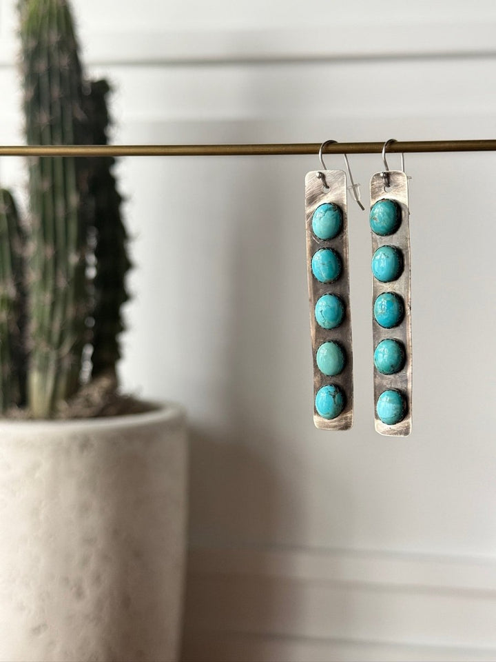 The Corrales Earrings by Savannah Sevens western life{&}style feature six round Kingman turquoise stones on each long, rectangular handmade earring, pictured hanging from a brass rod with a blurred cactus in the background.