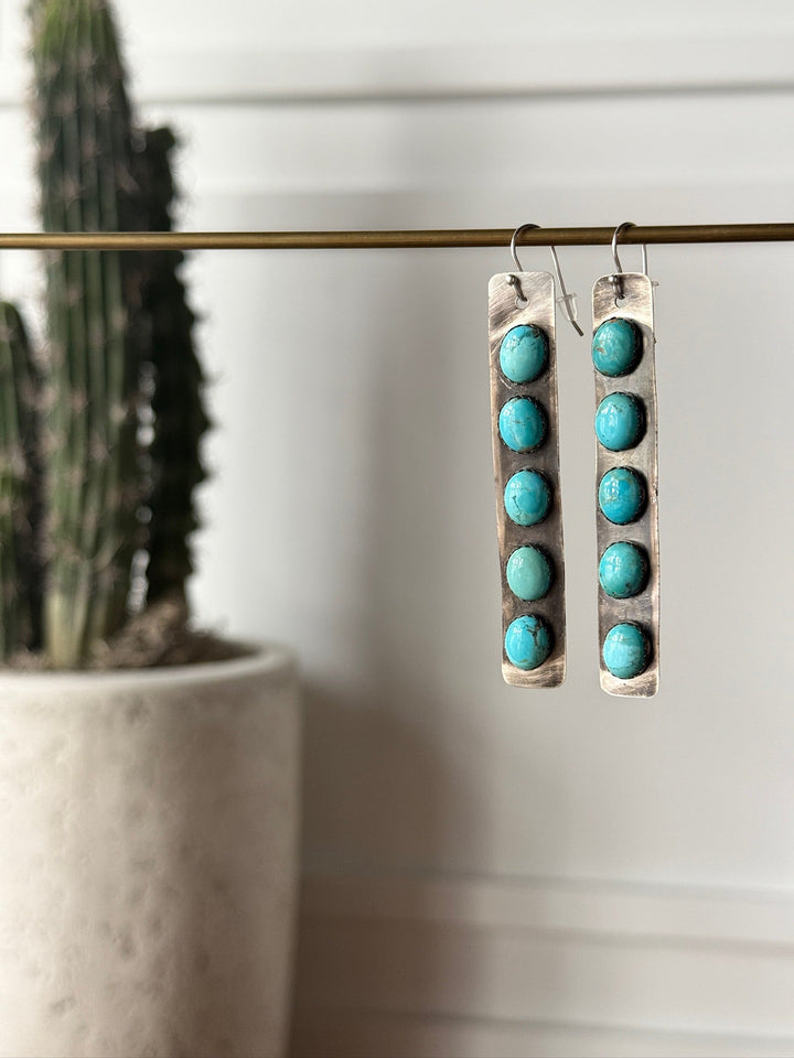The Corrales Earrings by Savannah Sevens western life{&}style feature six round Kingman turquoise stones on each long, rectangular handmade earring, pictured hanging from a brass rod with a blurred cactus in the background.