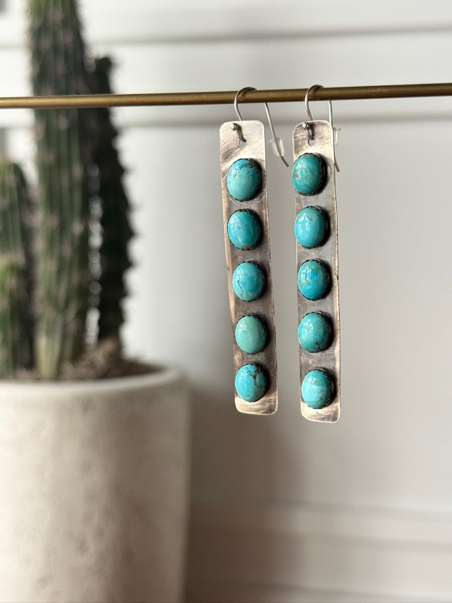 The Corrales Earrings by Savannah Sevens western life{&}style are long, rectangular sterling silver earrings featuring six round Kingman turquoise stones each, hanging from a rod with a blurred potted cactus in the background.
