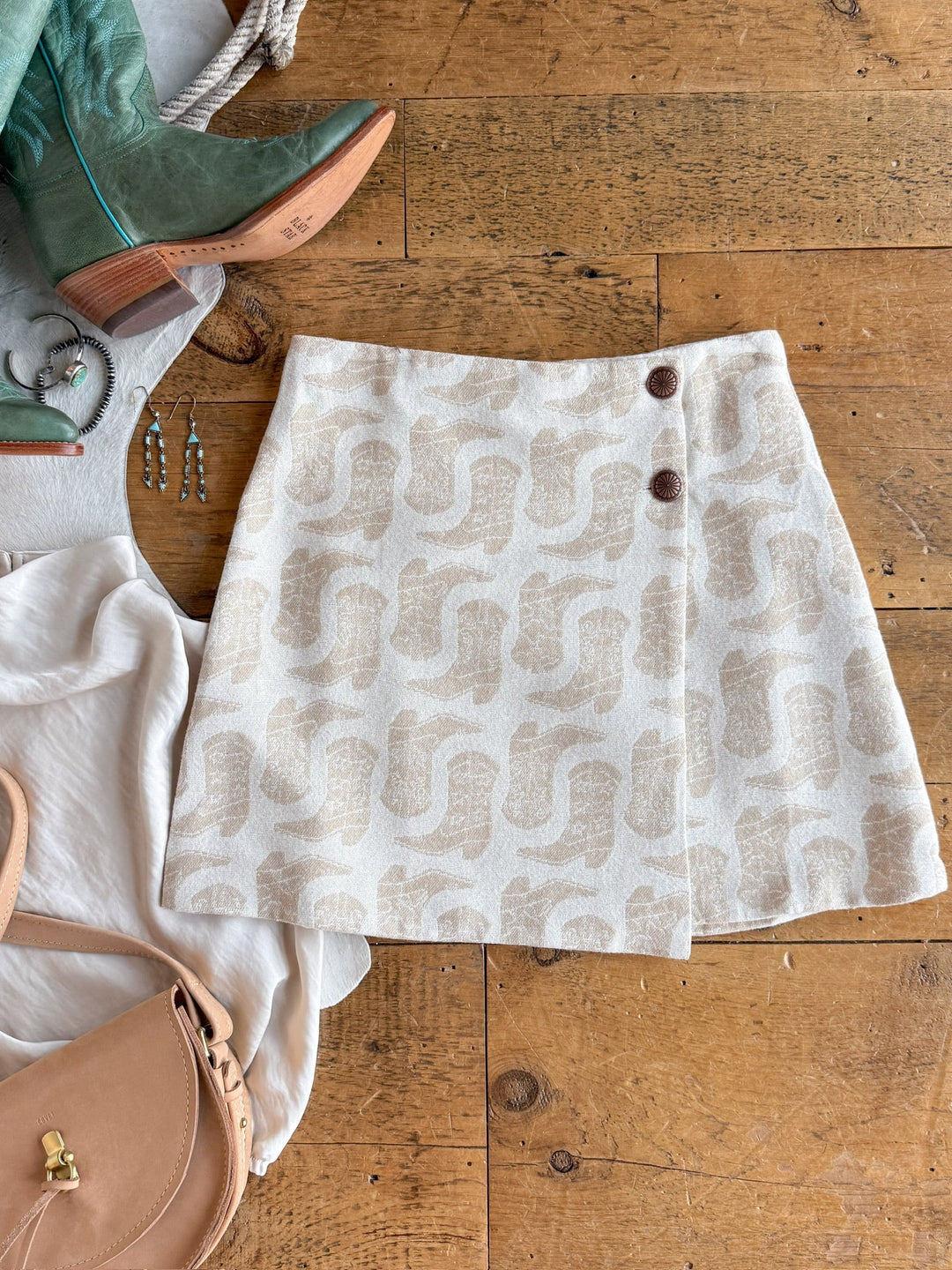 The Corsicana Cream Western Skirt by Savannah Sevens western life{&}style features a beige and cream pattern and high-waisted design with two brown concho buttons, displayed on a wooden floor alongside accessories including a green boot, jewelry, a cream blouse, and a beige purse.