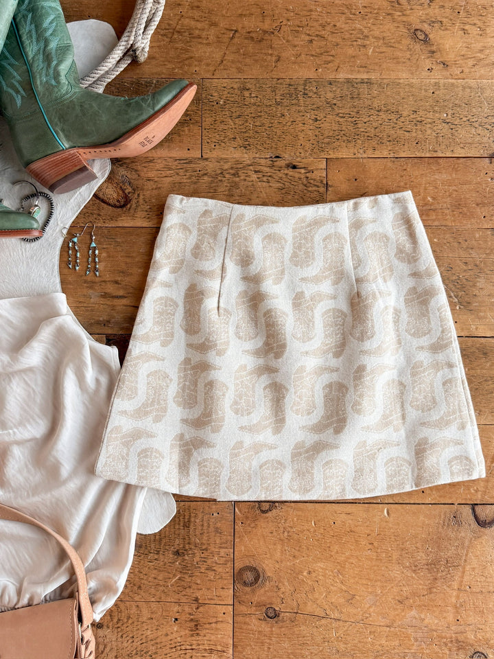 The Corsicana Cream Western Skirt by Savannah Sevens western life{&}style lies on a wooden floor next to a white blouse, tan sandals, turquoise boots, and a pair of earrings.