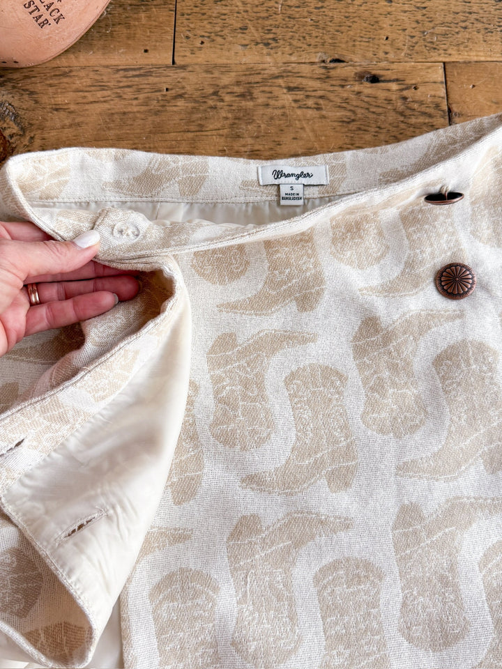 A hand displays the inside of the Corsicana Cream Western Skirt by Savannah Sevens western life{&}style, featuring a cream-colored cotton fabric with a squirrel pattern, a decorative brown concho button, and an inside snap closure, set on a wooden surface.