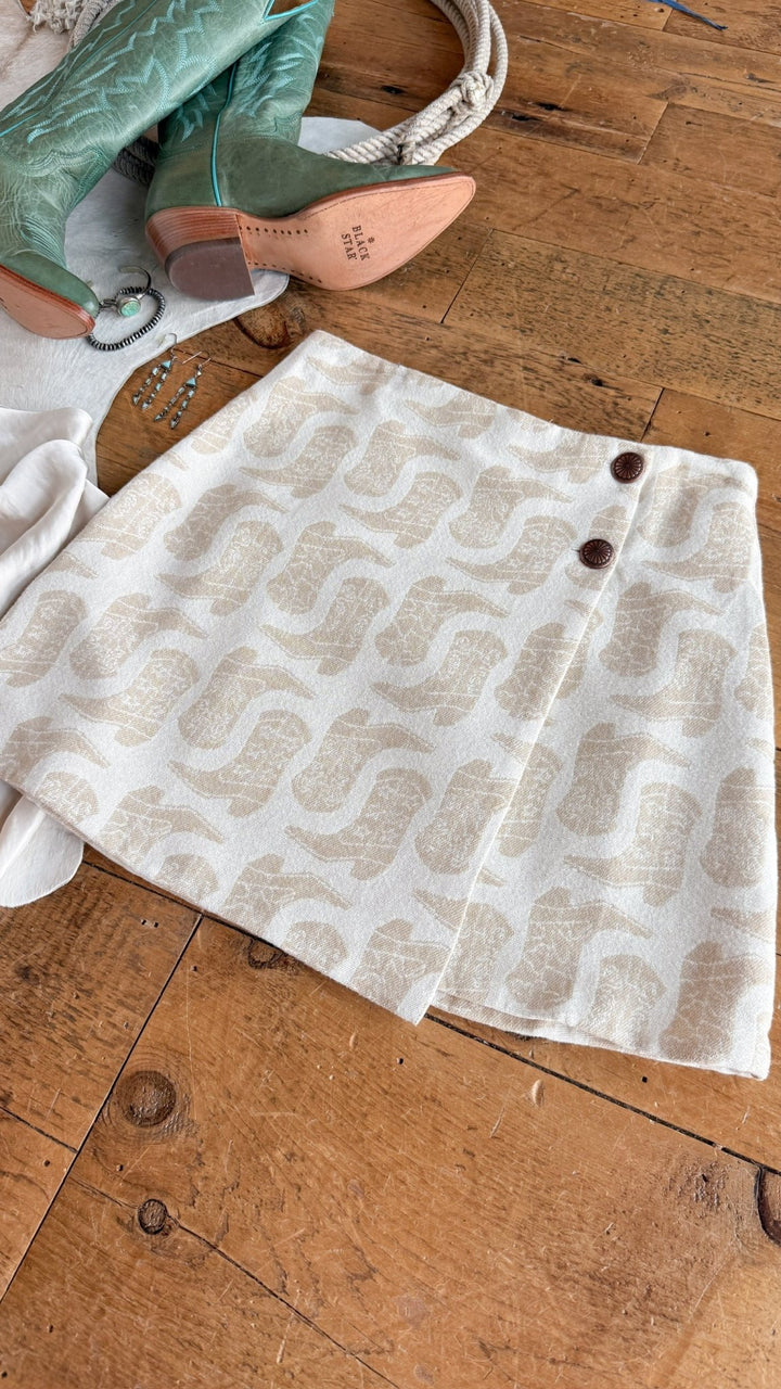 The Corsicana Cream Western Skirt by Savannah Sevens western life{&}style, featuring a beige and white pattern and two brown buttons at the high waist, is laid out on a wooden floor alongside green boots, a white purse, and silver jewelry.