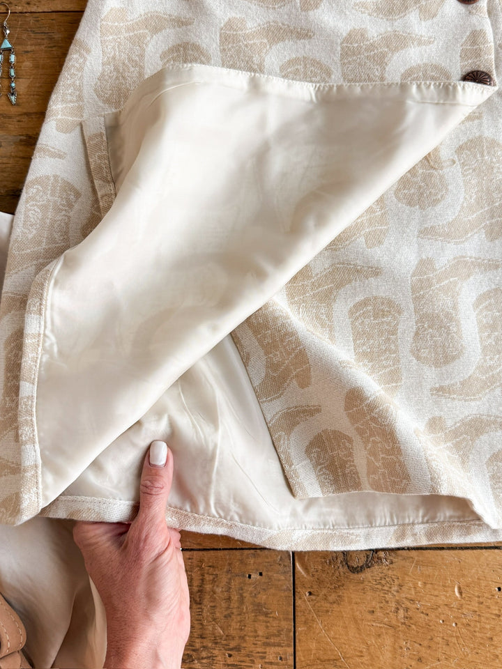 A hand lifts the cream-colored lining of the Corsicana Cream Western Skirt by Savannah Sevens western life{&}style, revealing its inner seam and cotton fabric texture. The skirt rests on a rustic wooden surface.