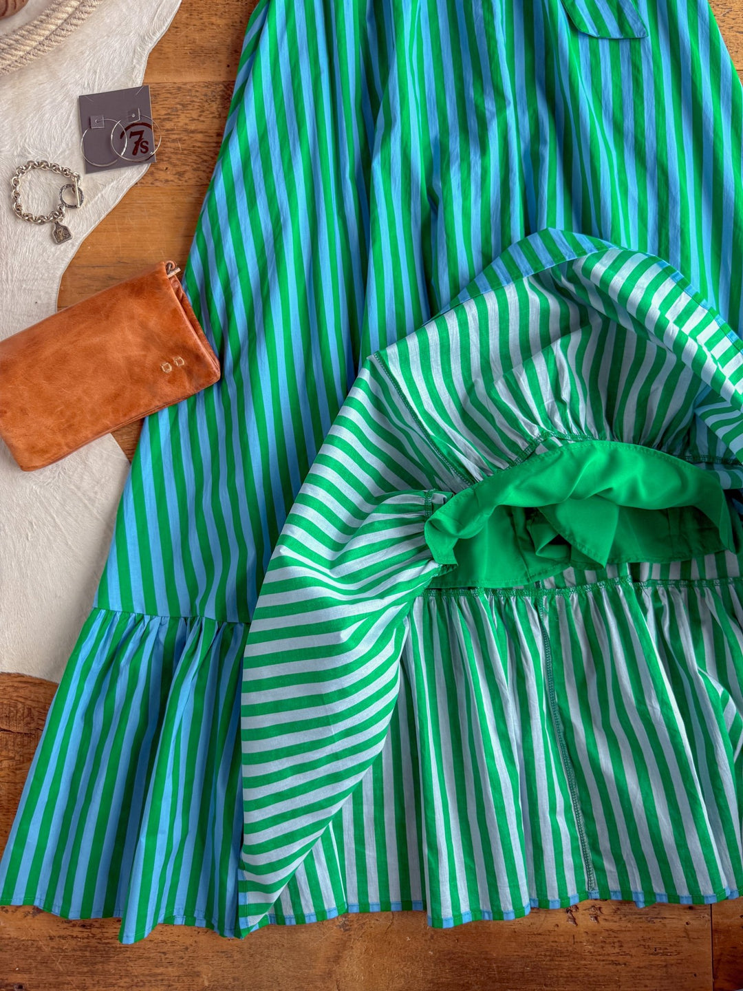 The Cortland Striped Halter Neck Dress by Savannah Sevens western life{&}style, featuring green and white stripes, a ruffled hem, and a green inner lining, is spread out on a wooden surface. Next to it are a brown clutch, a silver bracelet, and a pair of earrings.