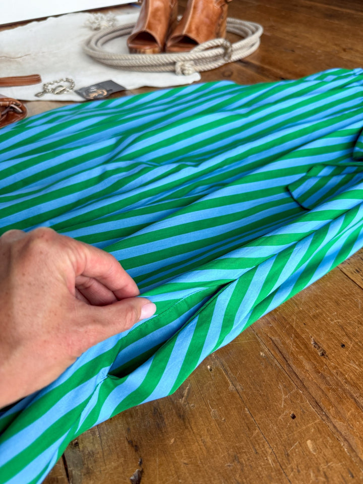 A hand holds the pocket of the Cortland Striped Halter Neck Dress by Savannah Sevens western life{&}style, featuring green and blue stripes and laid flat on a wooden floor, with brown shoes and accessories softly blurred in the background.