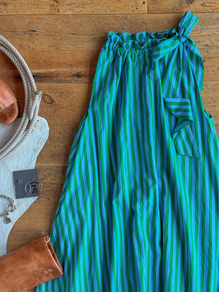 The Cortland Striped Halter Neck Dress by Savannah Sevens western life{&}style, featuring blue and green stripes and a bow at the waistband, is displayed on a wooden surface alongside a tan leather bag, white bag, keychain, earrings, and a rope handle.