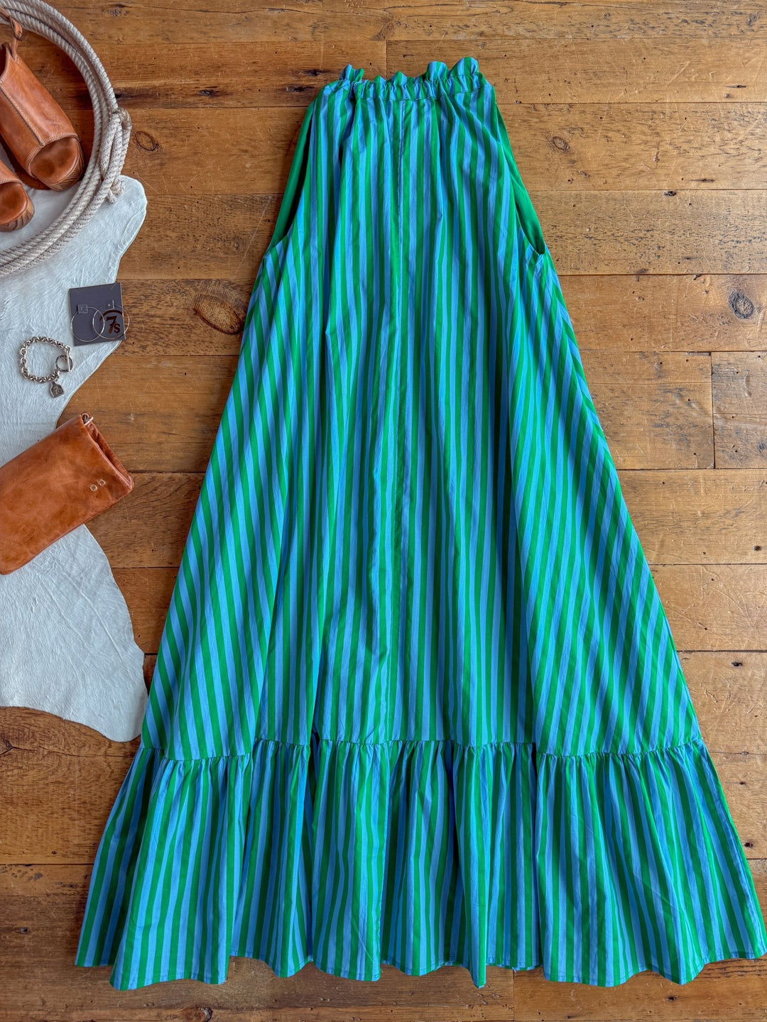 The Cortland Striped Halter Neck Dress by Savannah Sevens western life{&}style, featuring blue and green stripes with a ruffled hem, is laid out on a wooden floor alongside brown leather sandals, a bracelet, and a rust-colored clutch.