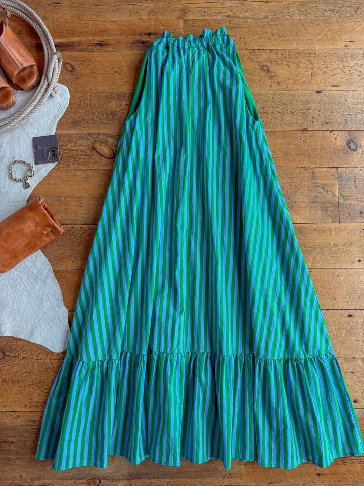 The Cortland Striped Halter Neck Dress by Savannah Sevens western life{&}style, featuring blue and green stripes with a ruffled hem, is laid out on a wooden floor alongside brown leather sandals, a bracelet, and a rust-colored clutch.