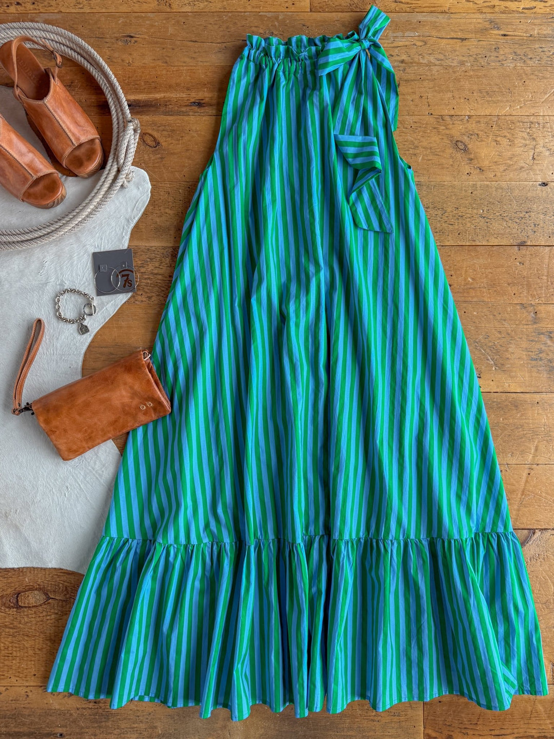 The Cortland Striped Halter Neck Dress by Savannah Sevens western life{&}style, featuring blue and green stripes and a ruffled hem, is displayed on a wooden floor. Beside it are tan heeled sandals, a brown clutch, a bracelet, and a white rug with rope detail.