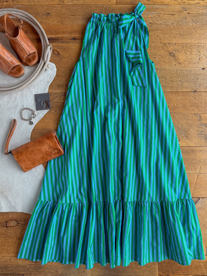 The Cortland Striped Halter Neck Dress by Savannah Sevens western life{&}style, featuring blue and green stripes and a ruffled hem, is displayed on a wooden floor. Beside it are tan heeled sandals, a brown clutch, a bracelet, and a white rug with rope detail.