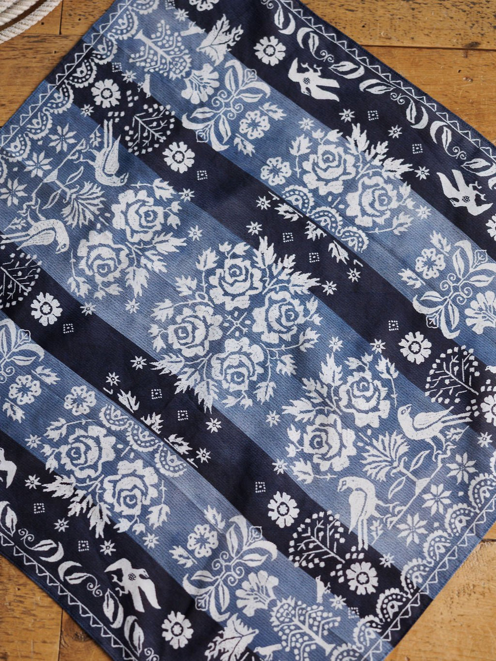The Coverlet Western Floral Cotton-Silk Neck Scarf by Savannah Sevens western life{&}style features blue and white floral designs, roses, birds, and geometric borders, displayed on a wooden surface.
