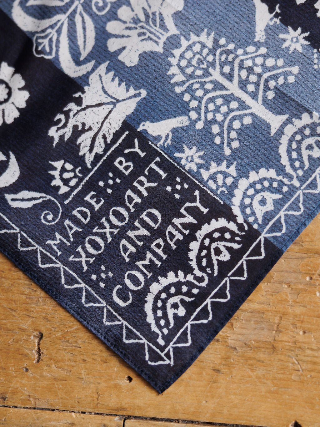 A close-up of the Coverlet Western Floral Cotton-Silk Neck Scarf by Savannah Sevens western life{&}style, featuring a blue and white pattern and a corner label, displayed on a wooden surface.